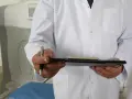 Lekarz w białym kitlu trzyma notatnik i długopis. Obok widoczny sprzęt medyczny, sugerujący badanie. Palpacja co to? To badanie fizykalne.