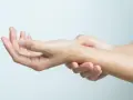 Dłoń z widocznym pieprzykiem na palcu, druga dłoń dotyka nadgarstka, gdzie wyczuwalna jest kość grochowata.