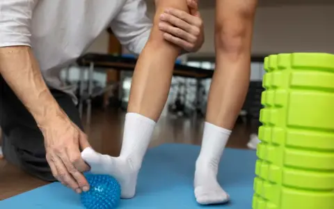 Fizjoterapeuta pomaga w rehabilitacji stawu skokowego, masując stopę pacjenta niebieską piłeczką. Obok stoi zielony wałek do masażu.