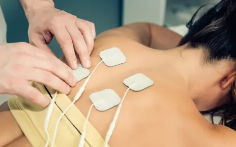 Fizjoterapeuta umieszcza elektrody na plecach pacjentki, przygotowując do zabiegu TENS. Należy pamiętać o przeciwwskazaniach do jego stosowania.