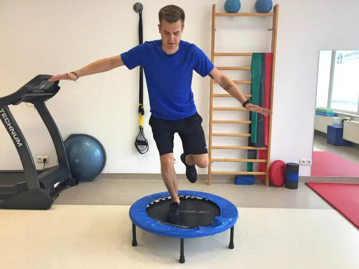 Mężczyzna wykonuje ćwiczenia po rekonstrukcji ACL, balansując na małej trampolinie. W tle bieżnia i drabinka gimnastyczna.