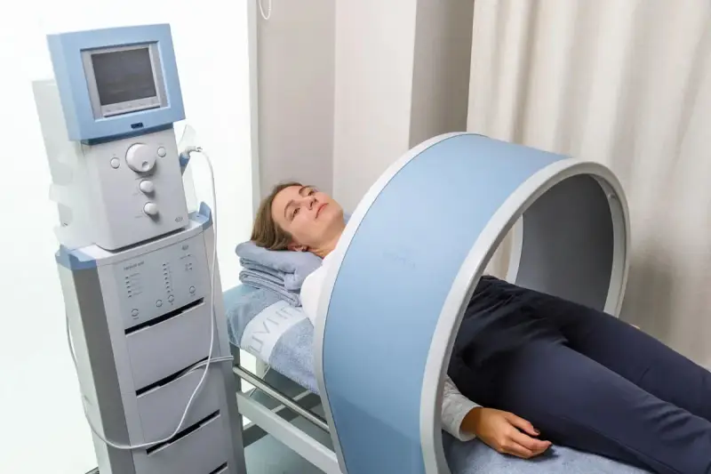 Kobieta leży na łóżku rehabilitacyjnym, poddając się zabiegowi z wykorzystaniem urządzenia generującego pole magnetyczne, które obejmuje jej kolano.